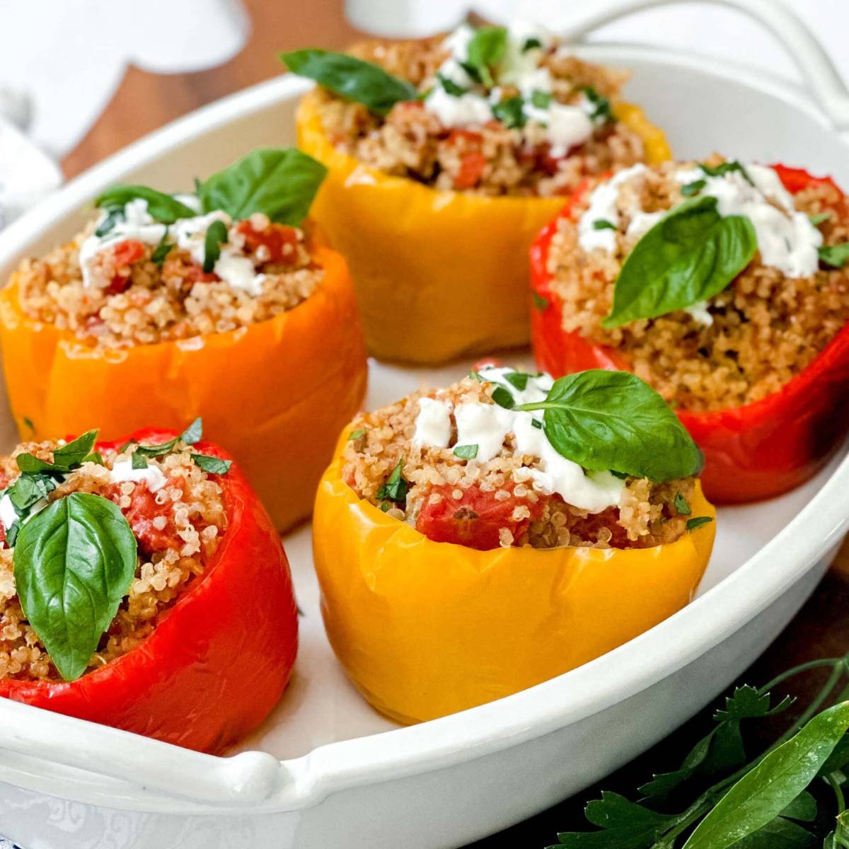 A white dish filled with stuffed peppers. The peppers are filled with a mixture of quinoa, tomatoes, and other vegetables. They are topped with a dollop of sour cream and fresh basil leaves.