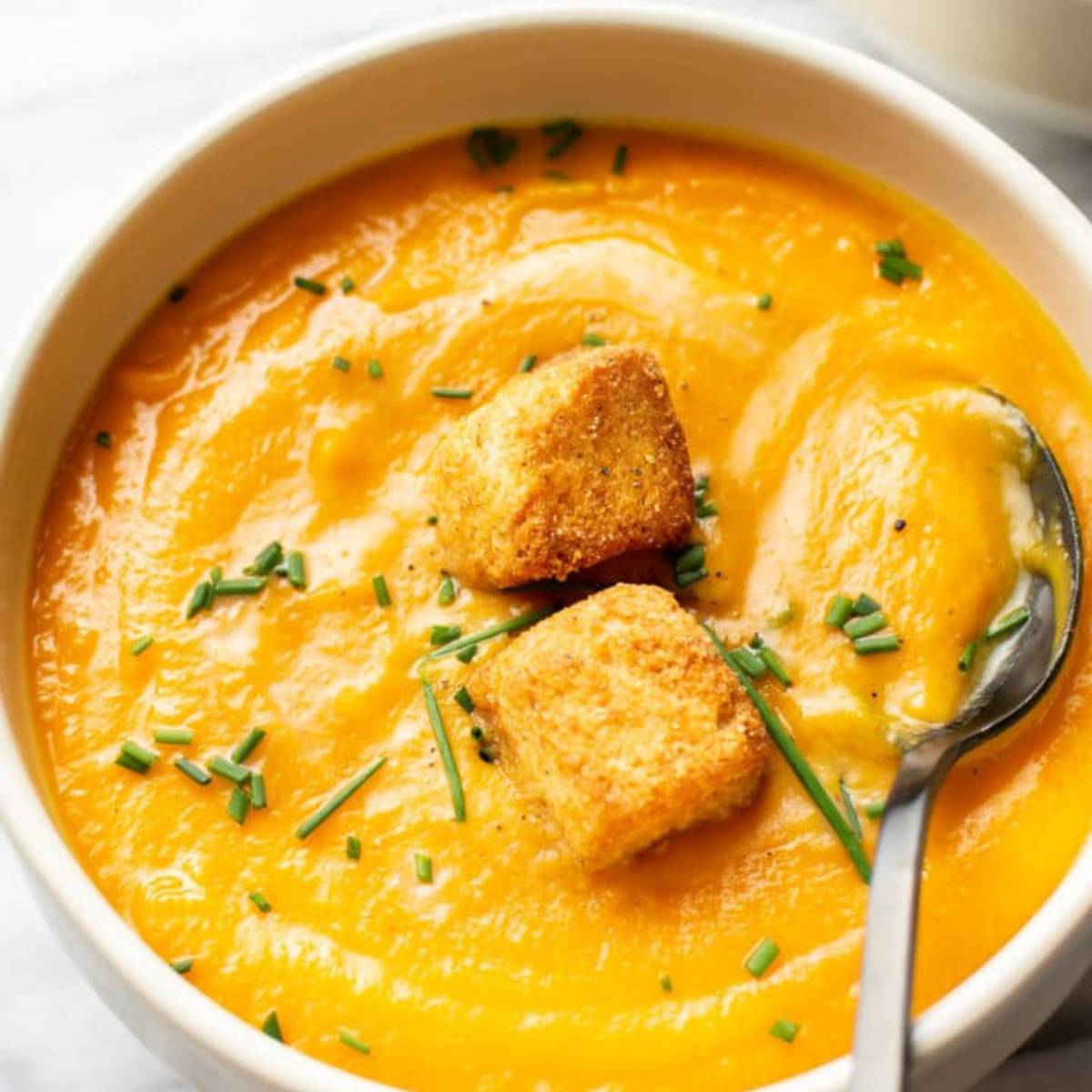  A bowl of creamy butternut squash soup, topped with croutons and chopped chives. The soup is a vibrant orange color.