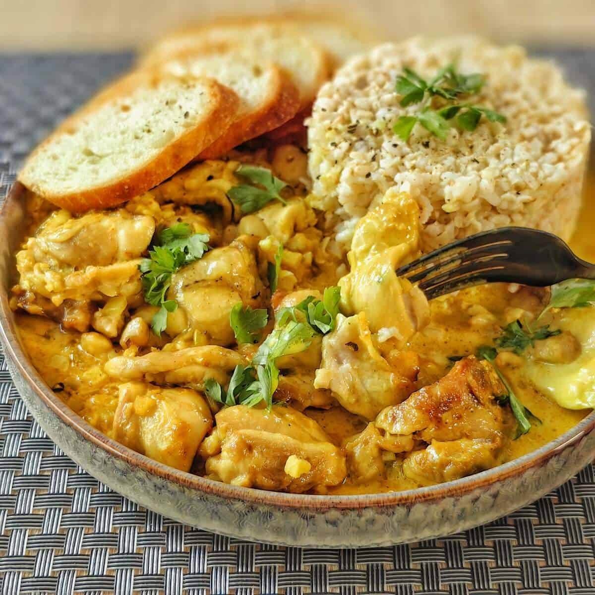 A plate of creamy chicken curry with a fork digging into it. The curry is served with a side of brown rice and toasted bread slices.