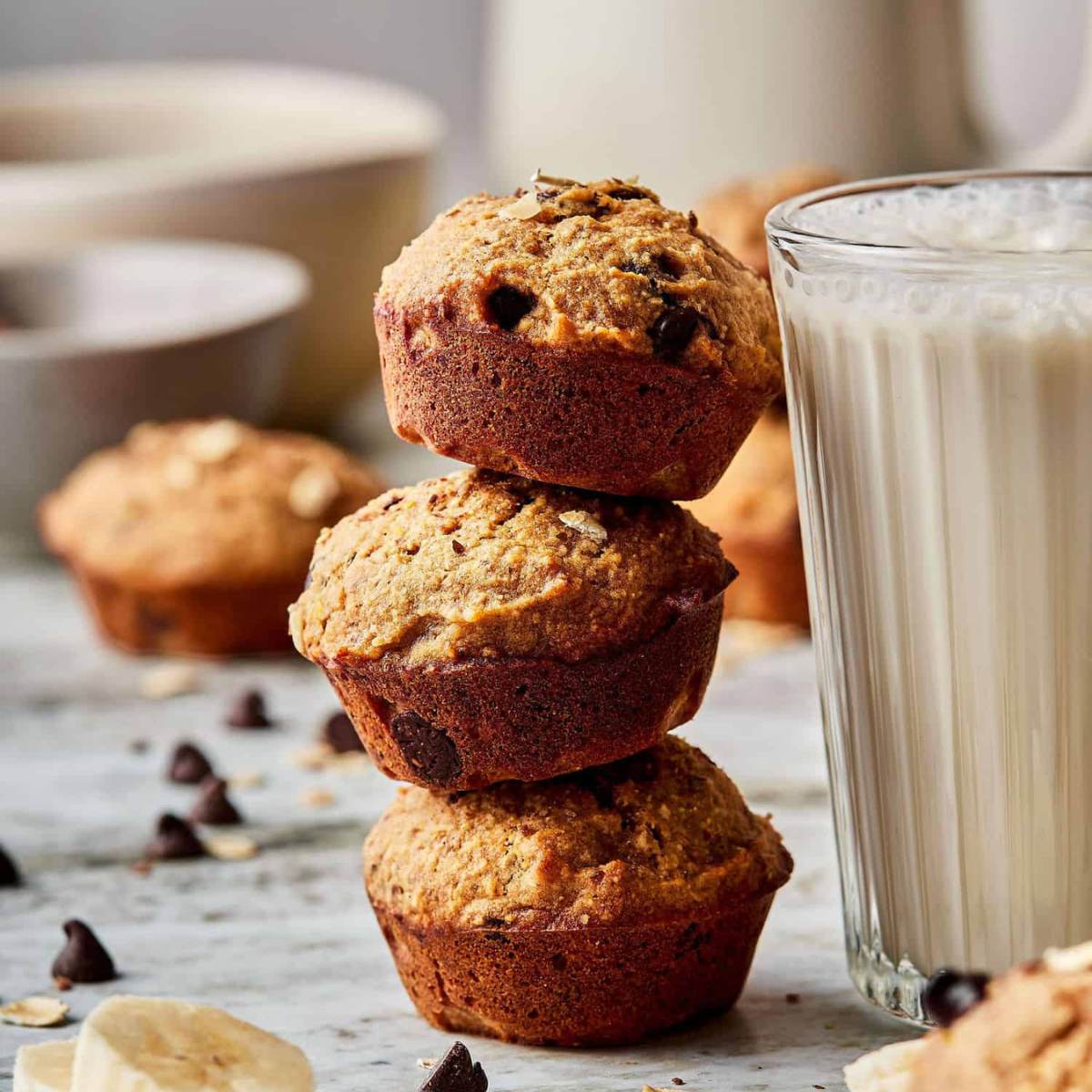 Three mini banana chocolate chip muffins stacked on top of each other, with a glass of milk and banana slices in the background.