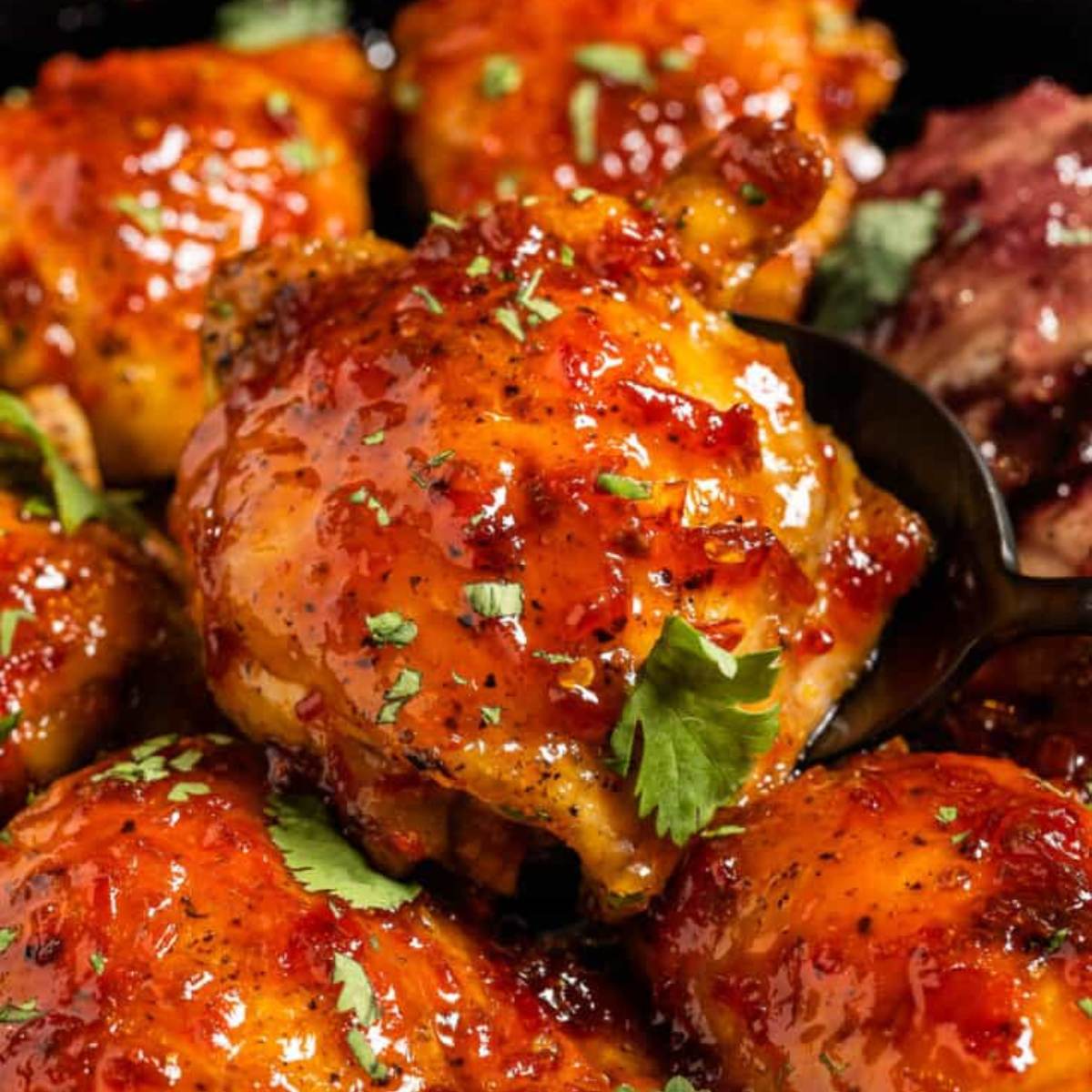  A close-up photo of a skillet filled with crispy, sweet and spicy chicken thighs. The chicken is glazed with a red sauce and garnished with fresh cilantro.
