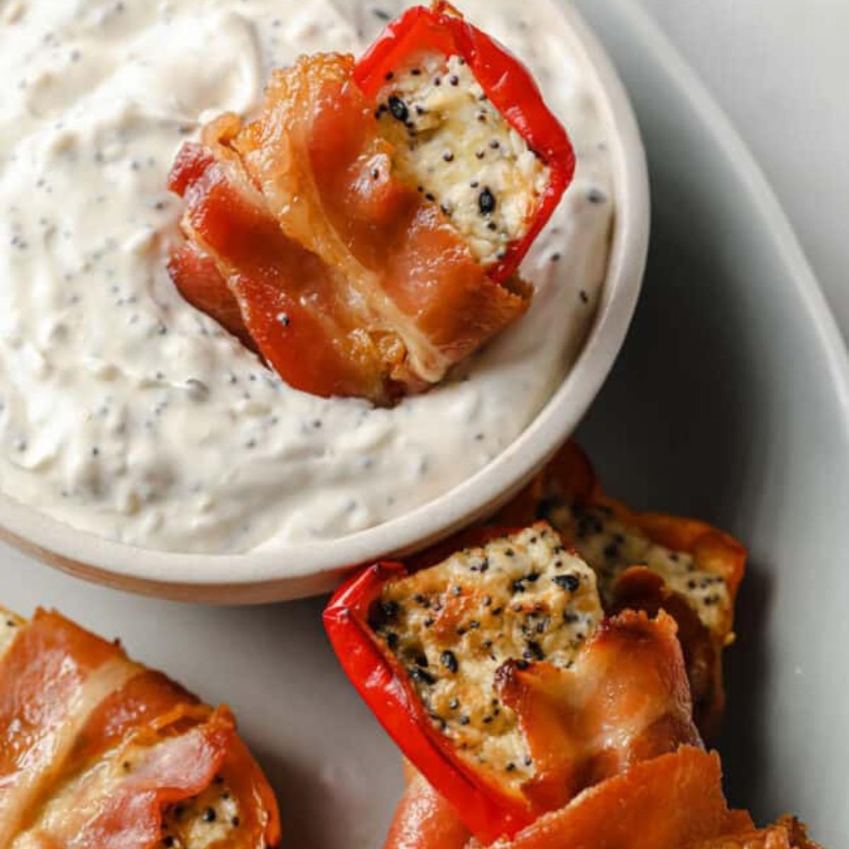 Bacon-wrapped mini peppers filled with a creamy cheese mixture, served with a small bowl of dipping sauce.