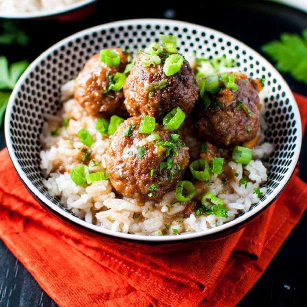 A bowl of sweet and sour meatballs served over a bed of white rice. The meatballs are glazed in a tangy sauce and garnished with chopped green onions and fresh parsley.