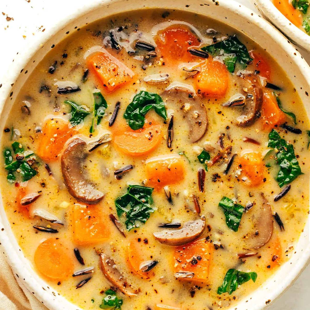 A bowl of creamy wild rice soup with mushrooms, carrots, and kale. The soup is a light brown color and is topped with fresh herbs.