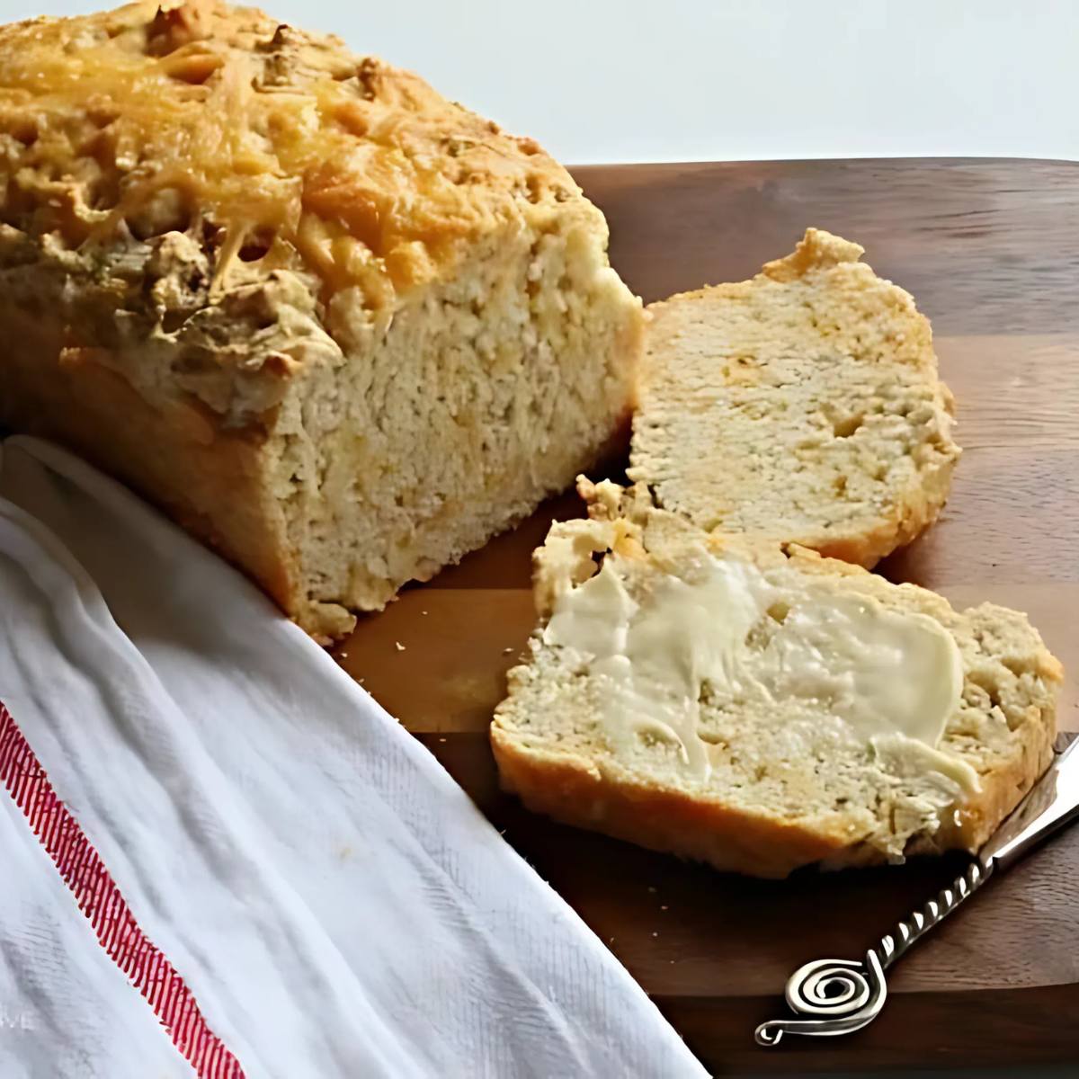 A golden-brown loaf of cheese bread, sliced on a wooden cutting board, with a piece of butter melting on one slice.