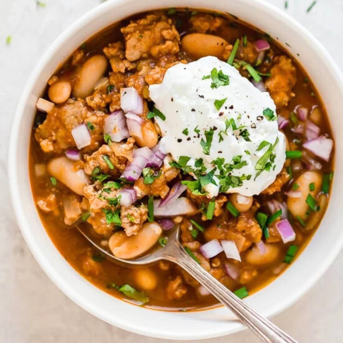 A bowl of hearty turkey chili, filled with ground turkey, white beans, tomatoes, and onions. The chili is topped with a dollop of sour cream, chopped green onions, and fresh parsley.