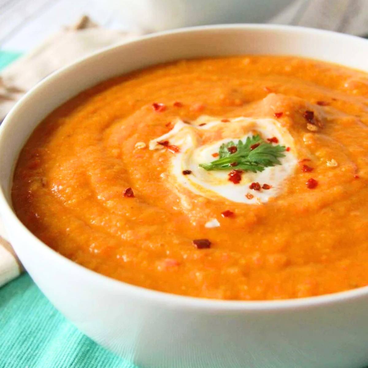 A bowl of creamy carrot soup, topped with a dollop of yogurt, red pepper flakes, and fresh cilantro. The soup is a vibrant orange color and is served in a white bowl on a turquoise napkin.