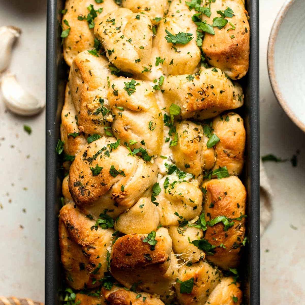 A golden-brown pull-apart bread loaf, filled with melted cheese and herbs, in a loaf pan.