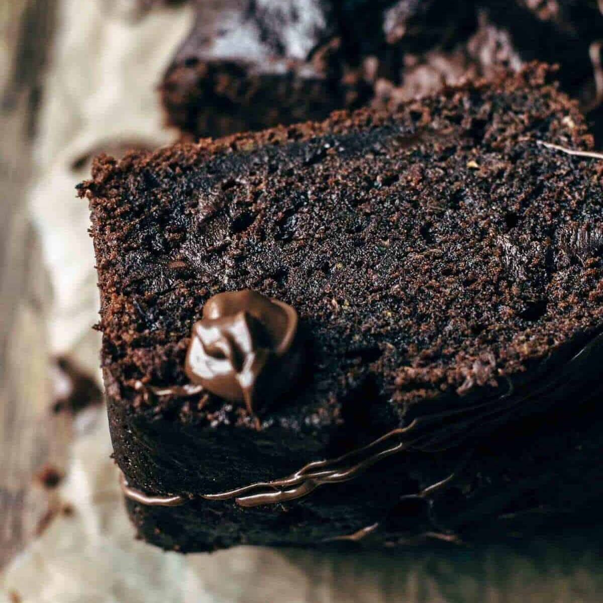 A close-up of a slice of dark chocolate zucchini bread, drizzled with chocolate and topped with a small chocolate square.