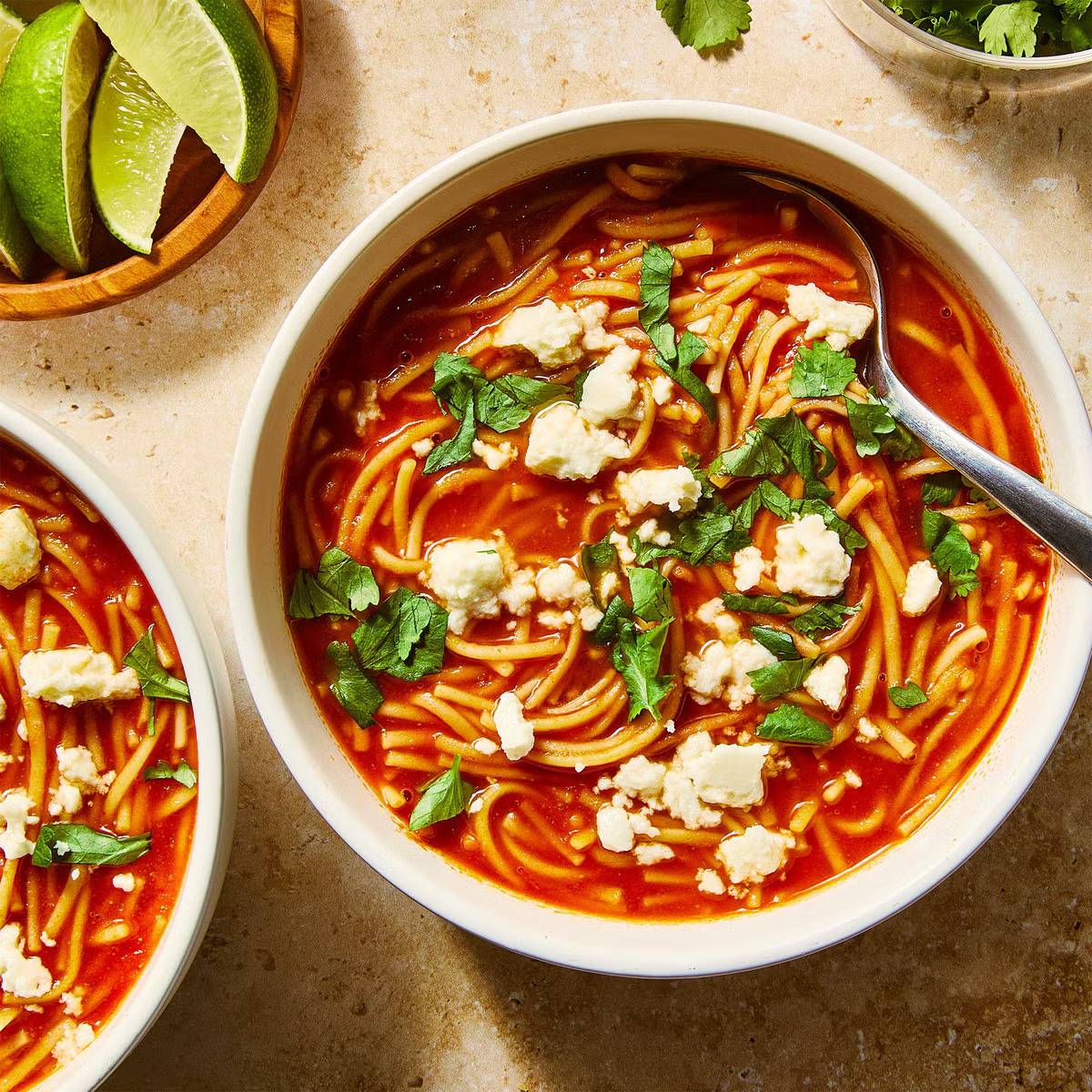 Two bowls of noodle soup. The soup is a vibrant red color and contains thin noodles, shredded cheese, and fresh cilantro. 