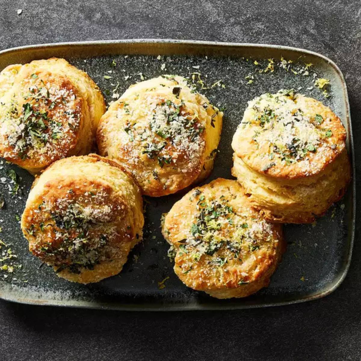 A platter of five golden-brown biscuits, topped with a mixture of lemon zest, herbs, and cheese.