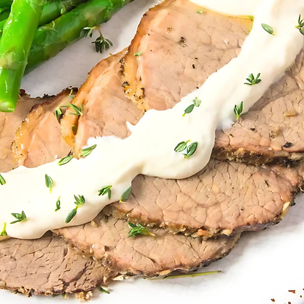 A plate of sliced roast beef, topped with a creamy horseradish sauce and fresh thyme. The beef is served with a side of roasted asparagus.