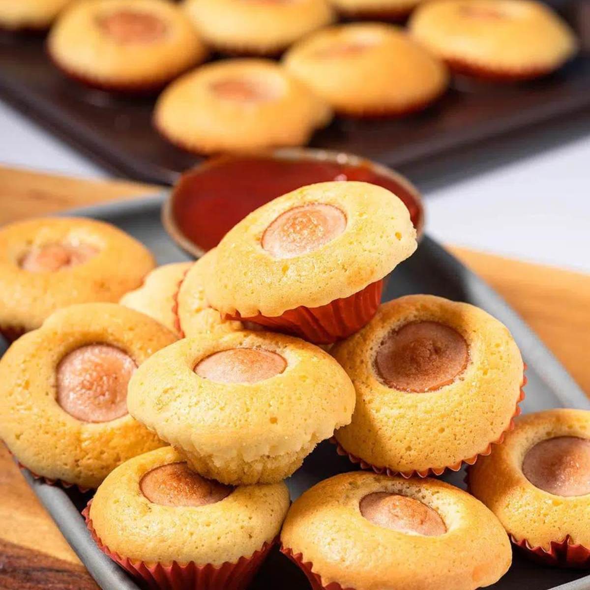 A pile of mini corn dog muffins, with a small bowl of ketchup and a muffin tin in the background.