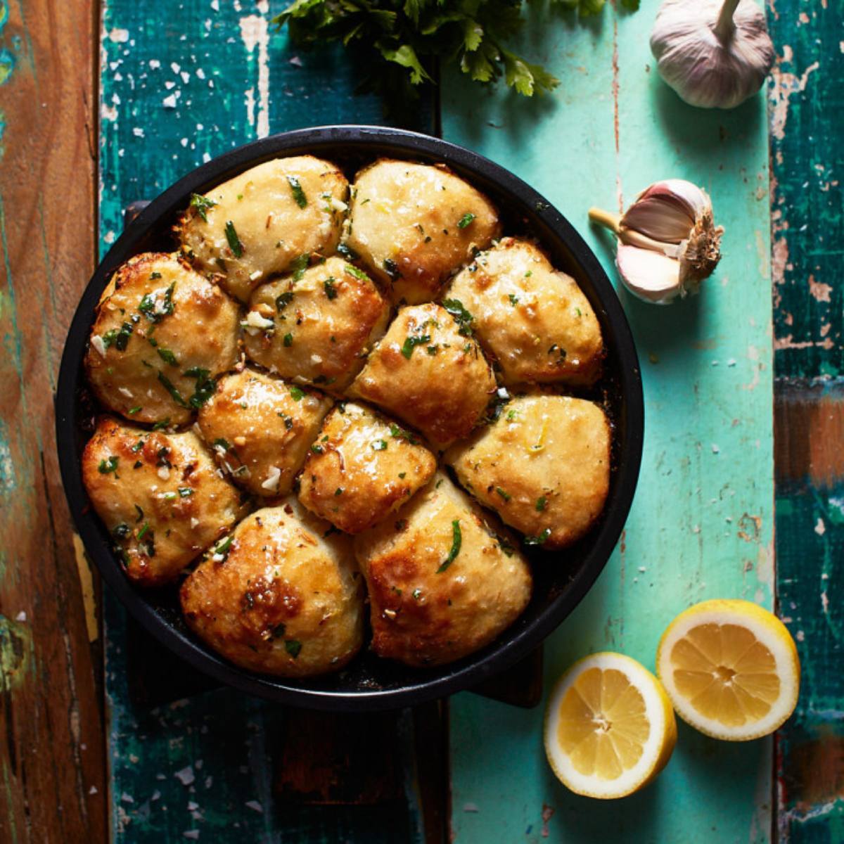 A cast iron skillet filled with golden-brown dinner rolls, topped with melted butter, garlic, herbs, and lemon slices.