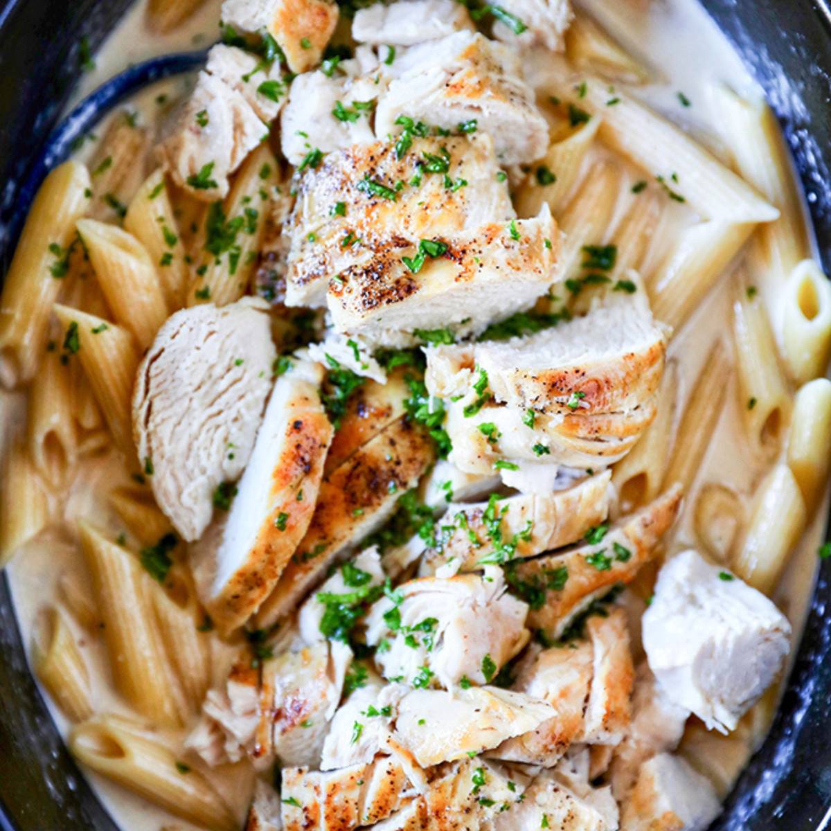  A close-up photo of a crockpot filled with penne pasta and shredded chicken in a creamy Alfredo sauce. Fresh parsley is sprinkled on top.