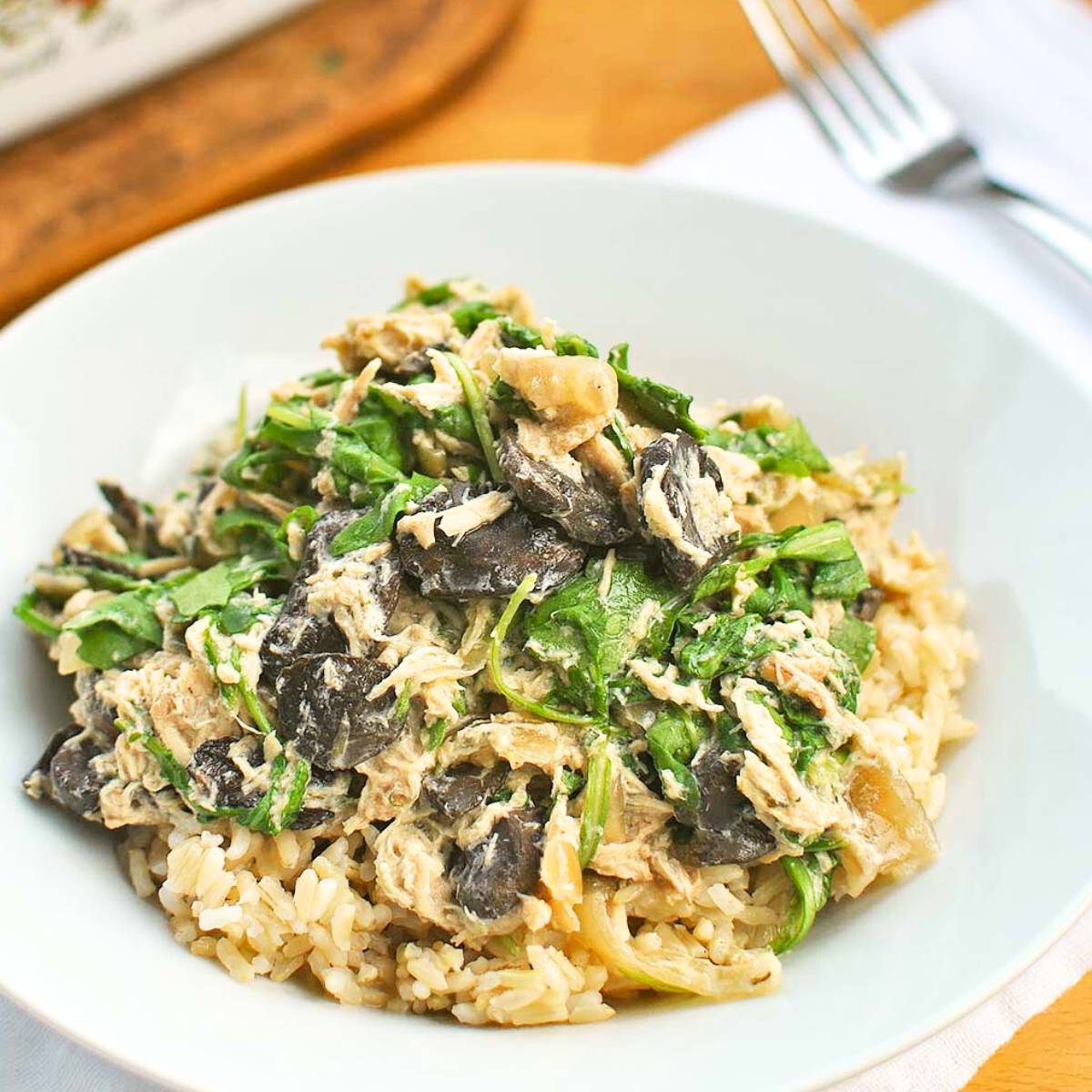 A plate of creamy mushroom and spinach chicken over brown rice. The chicken is shredded and coated in a creamy sauce, and the mushrooms are sautéed. There are fresh spinach leaves scattered throughout the dish.