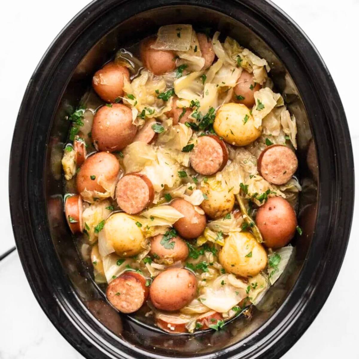 A slow cooker filled with a hearty stew made with cabbage, potatoes, and sausage. The vegetables are tender and the sausage is cooked through. The stew is topped with fresh parsley.