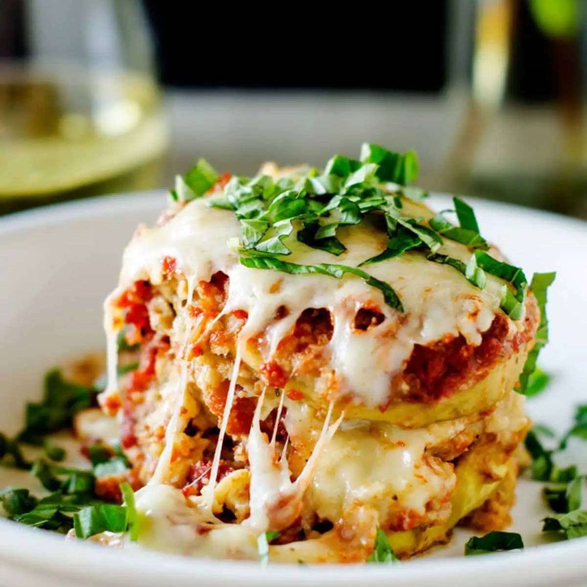 A plate of eggplant parmesan, with layers of eggplant, marinara sauce, and melted mozzarella cheese. The dish is topped with fresh basil leaves.