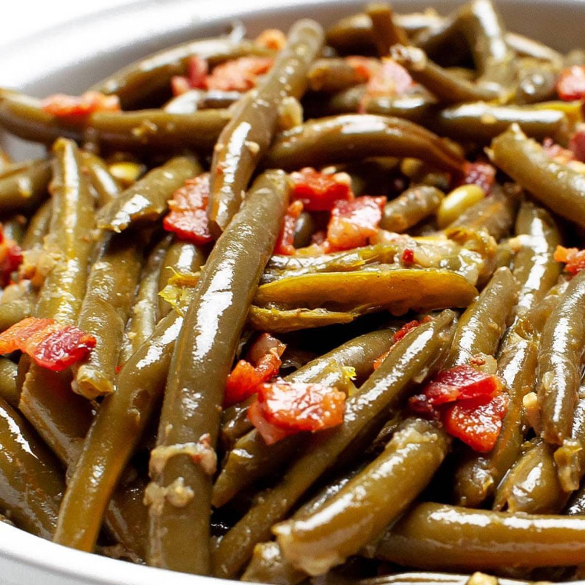 A close-up of a dish of green beans cooked with bacon. The green beans are bright green and coated in a glossy sauce, with bits of crispy bacon throughout.