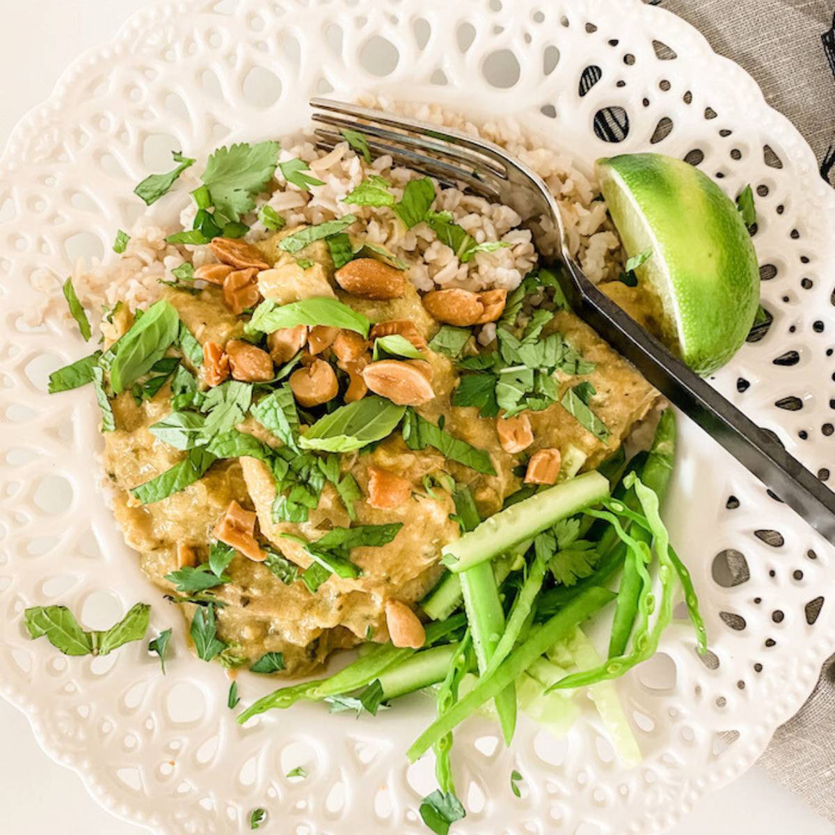 A plate of Thai green curry with chicken, served over brown rice. The curry is a vibrant green color and is topped with chopped peanuts, fresh cilantro, and a lime wedge. There are also cucumber slices on the side.