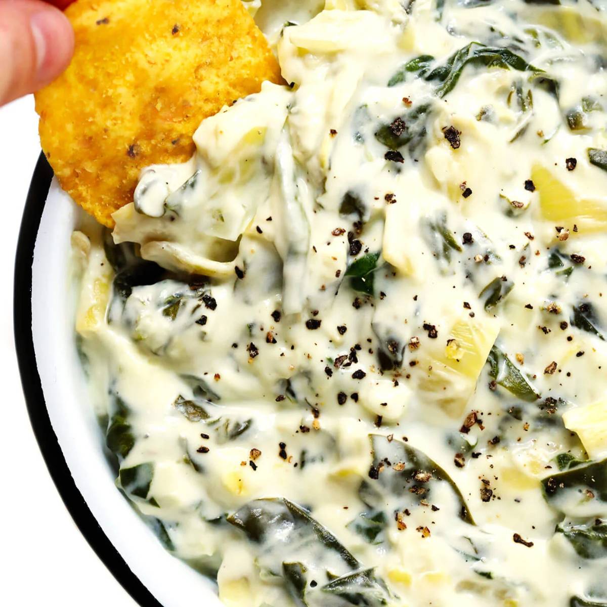 A bowl of creamy spinach artichoke dip, with a tortilla chip dipping into it. The dip is a pale green color and is topped with black pepper.