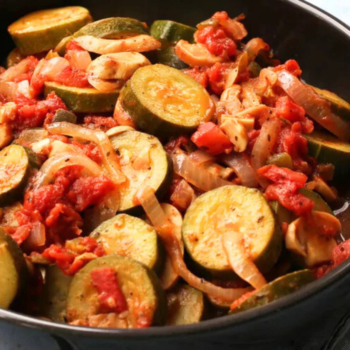 A skillet filled with sautéed zucchini, tomatoes, onions, and mushrooms. The vegetables are coated in a red sauce and are sizzling in the pan.