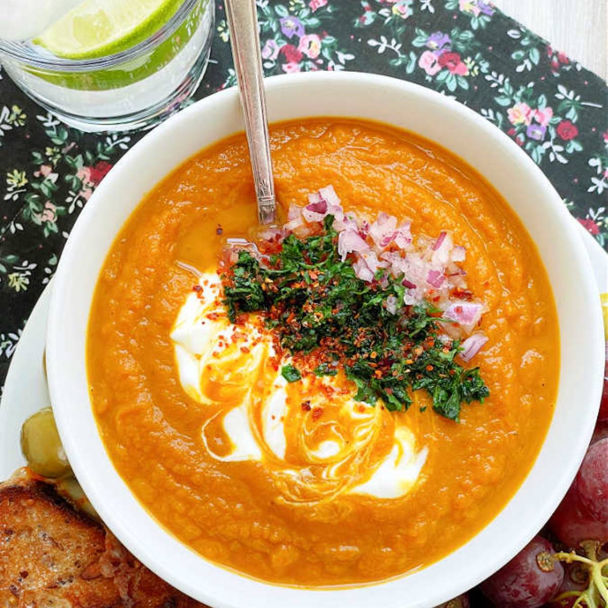 A bowl of creamy carrot soup topped with dollops of sour cream, chopped red onion, chopped parsley, and red pepper flakes.