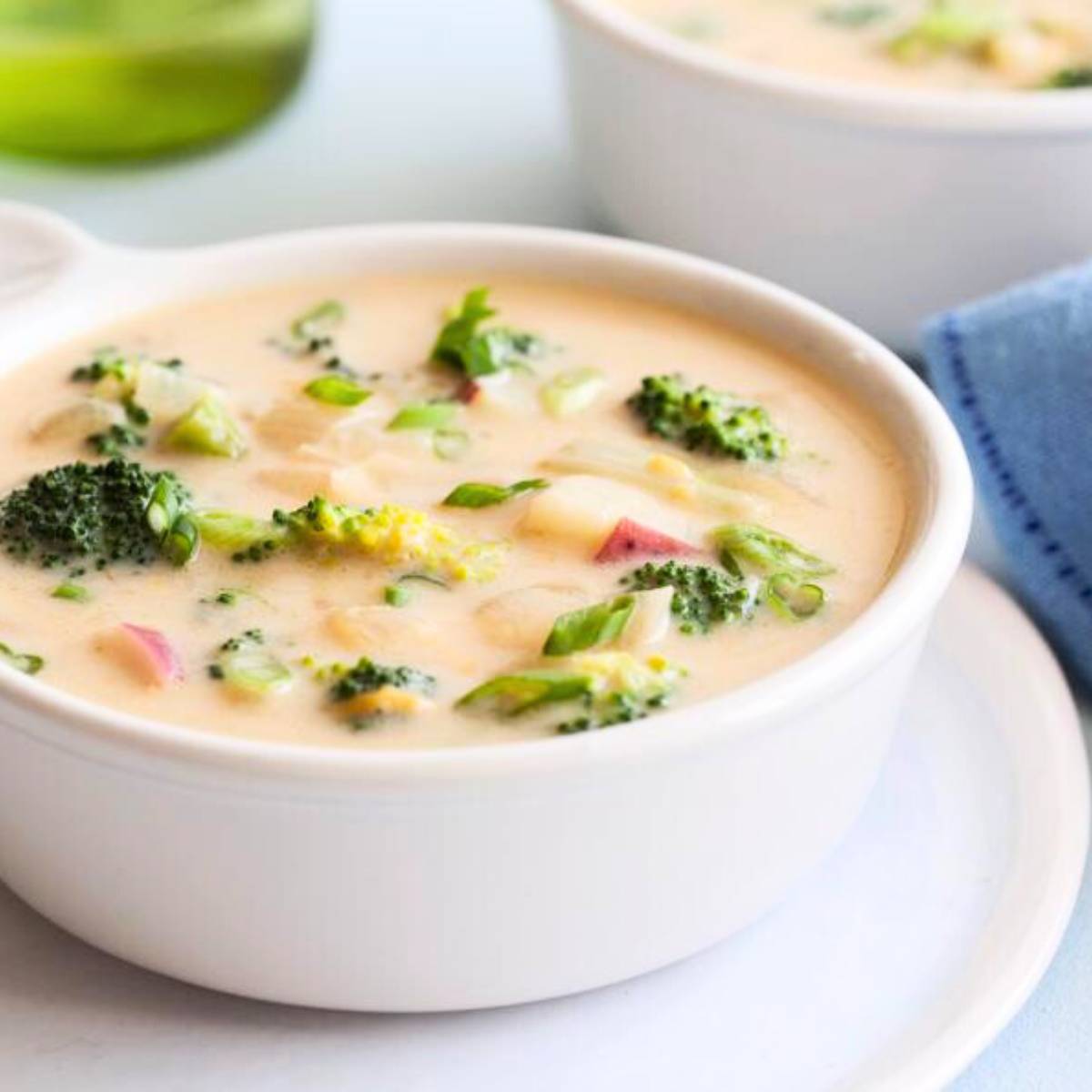 A close-up of a bowl of creamy broccoli cheddar soup, with visible chunks of broccoli, potatoes, and green onions. The soup is in a white bowl on a white plate, with a blue napkin and a wine bottle in the background.