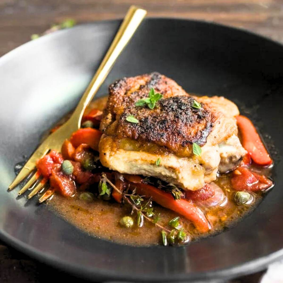 A plate of seared chicken thighs served with a red pepper sauce and capers. A gold-colored fork rests on the plate.