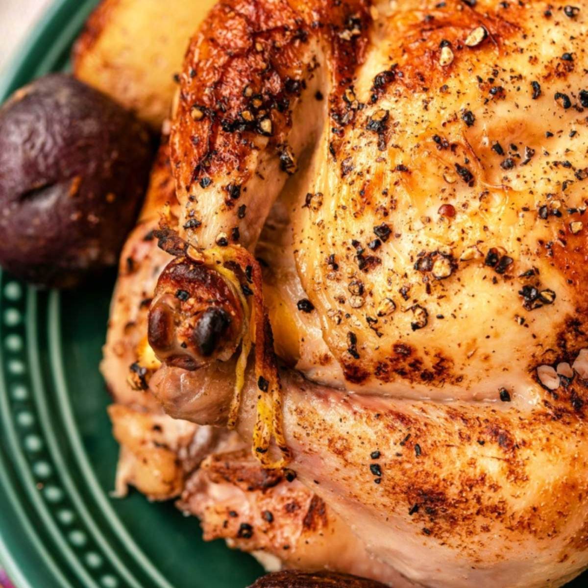  A close-up photo of a roasted whole chicken with crispy skin, seasoned with black pepper and herbs. There are roasted potatoes on the side.