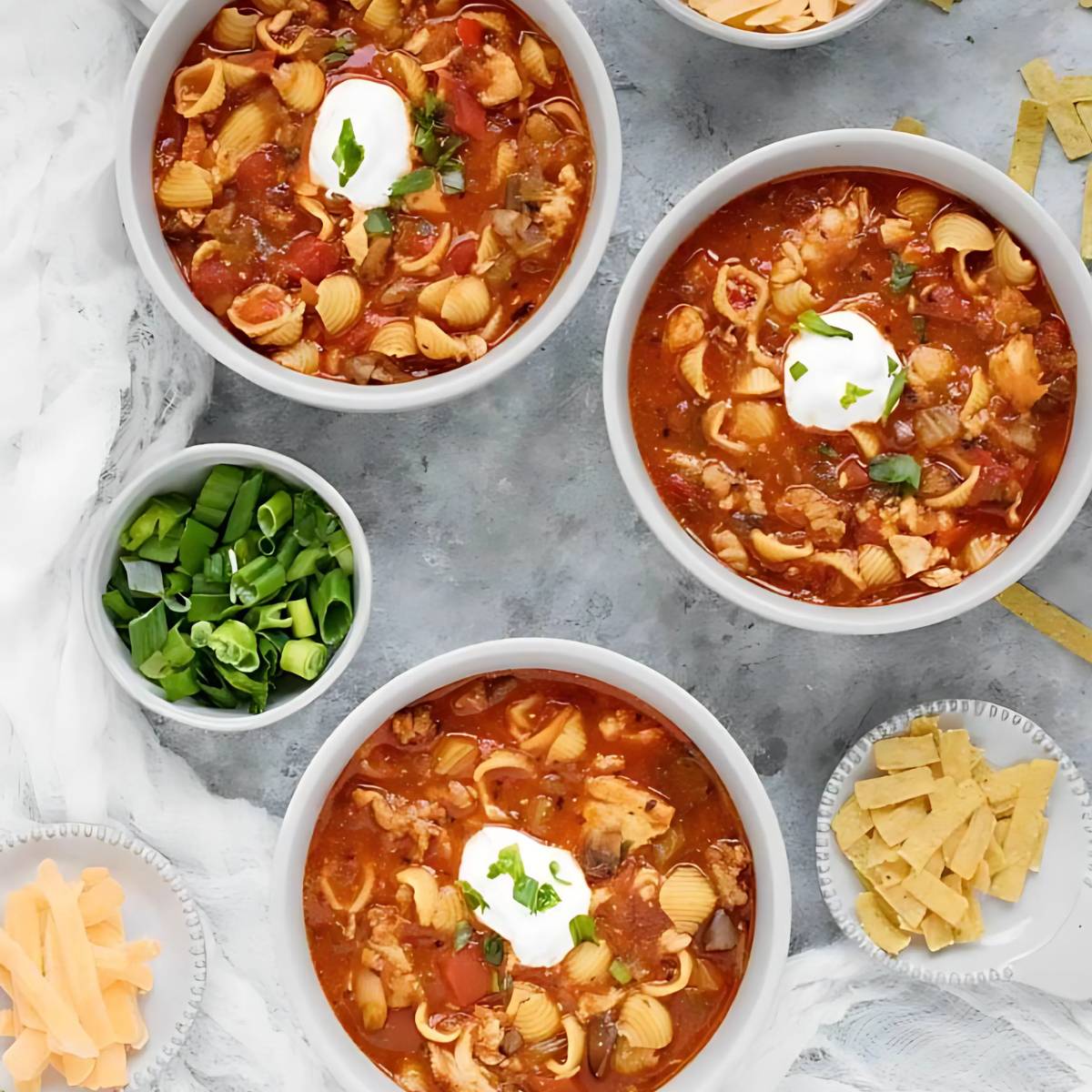 Three bowls of hearty chicken tortilla soup, topped with sour cream, shredded cheese, and chopped green onions. The soup contains pasta, vegetables, and chicken. 