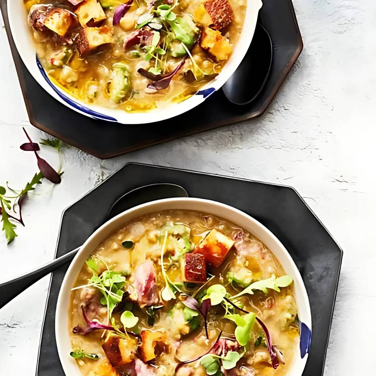 Two bowls of hearty smoked ham hock and lentil soup, topped with fresh herbs and croutons. The soup is a creamy, light brown color and contains visible chunks of ham hock and lentils.