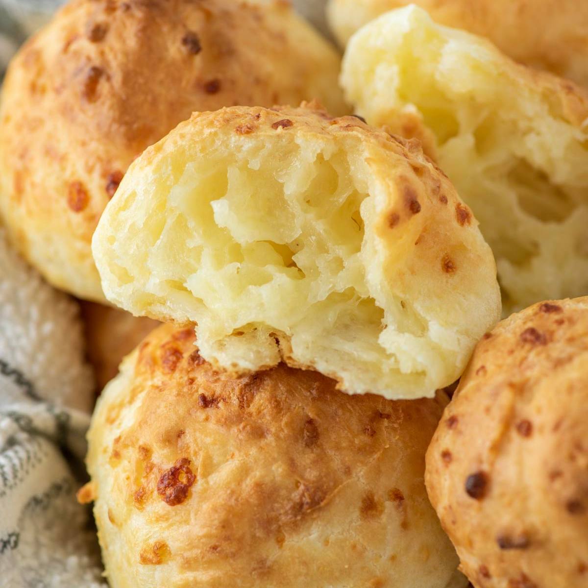 A close-up of golden-brown cheese bread rolls, one of which is broken open to reveal a soft, cheesy interior.