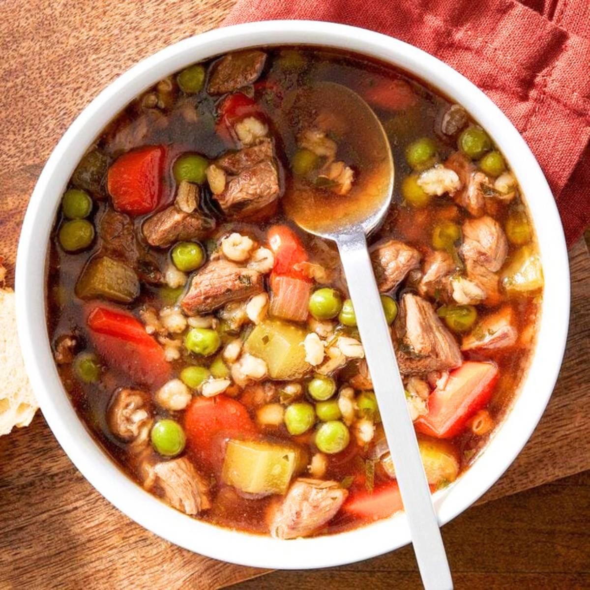 A bowl of hearty beef barley soup, filled with chunks of beef, barley, vegetables, and peas. A spoon rests in the bowl, and there's a slice of bread on the side.