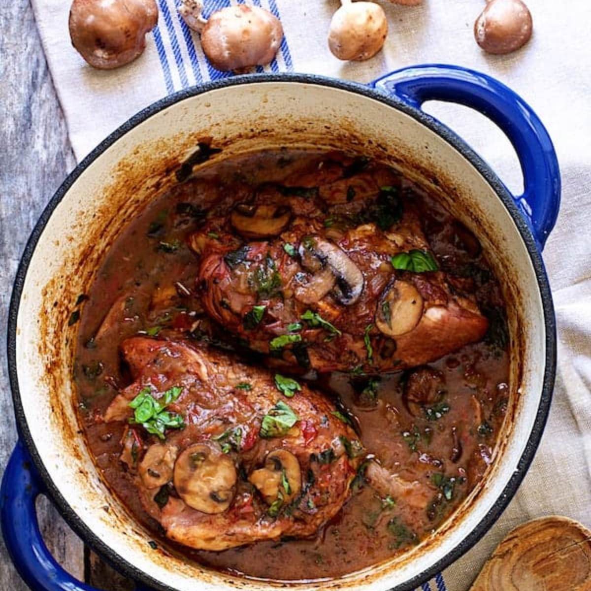  A blue Dutch oven filled with chicken smothered in a rich, mushroom sauce. Fresh basil leaves are scattered on top.