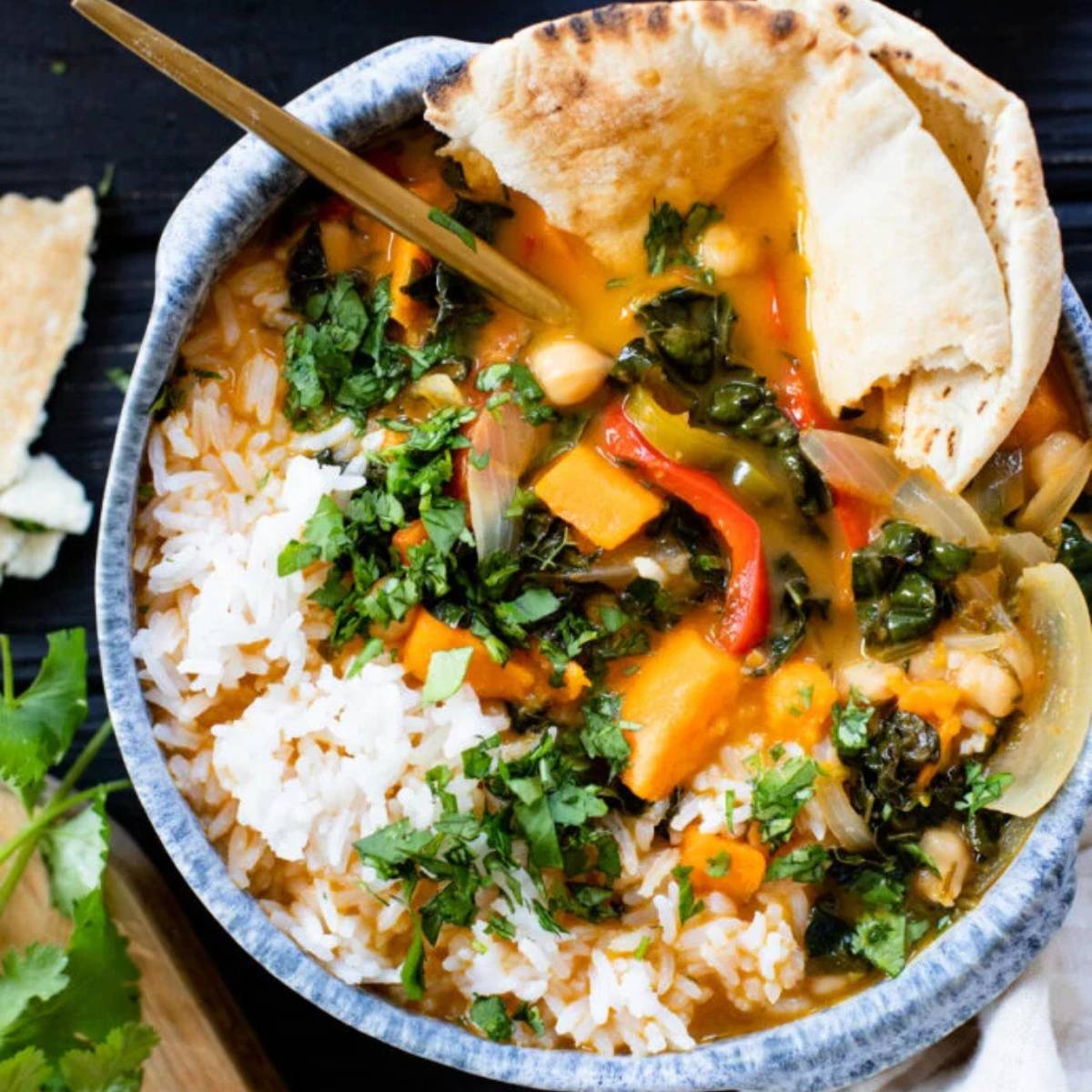  A bowl of Thai red curry with chickpeas, sweet potatoes, red bell peppers, and kale. It's served with a side of white rice and garnished with fresh cilantro. A piece of pita bread is resting on the side of the bowl.