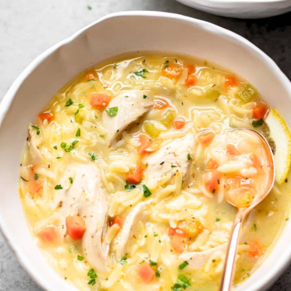 A bowl of creamy chicken orzo soup with lemon, filled with orzo pasta, shredded chicken, vegetables, and a lemon wedge. The soup is a pale yellow color and is topped with fresh parsley.