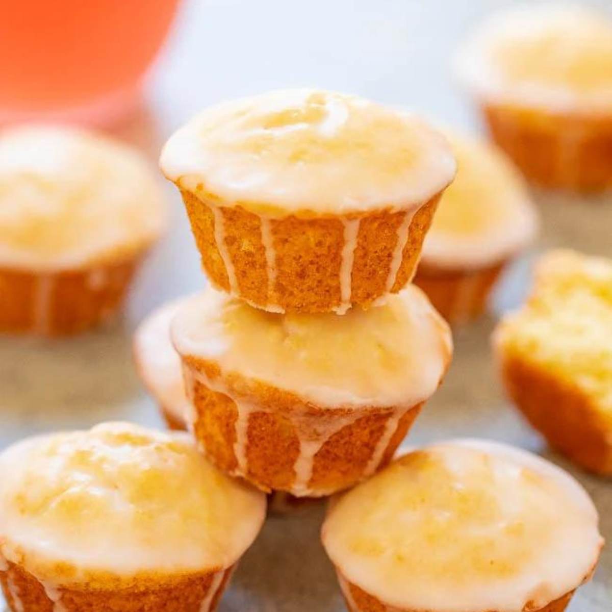 A stack of three mini orange glaze muffins, with additional muffins and a glass of orange juice in the background.