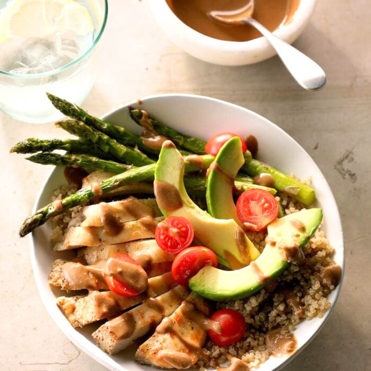  A bowl filled with grilled chicken, quinoa, roasted asparagus, cherry tomatoes, and avocado slices. A small bowl of peanut sauce is on the side.