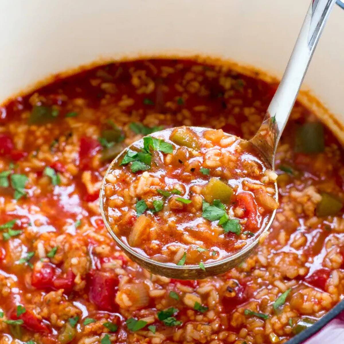 A ladle full of hearty sausage and rice soup, with visible chunks of sausage, rice, and vegetables. The soup is a vibrant red color and is served in a large pot.