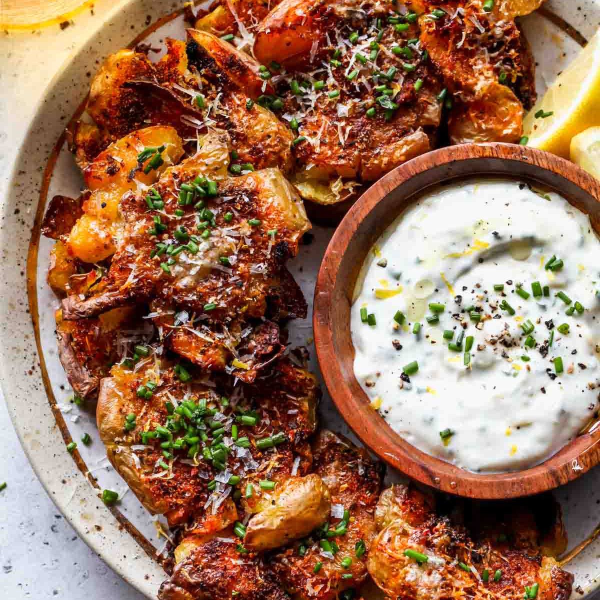 A plate of crispy, golden-brown smashed potatoes topped with chives and Parmesan cheese, served with a small bowl of creamy dipping sauce.