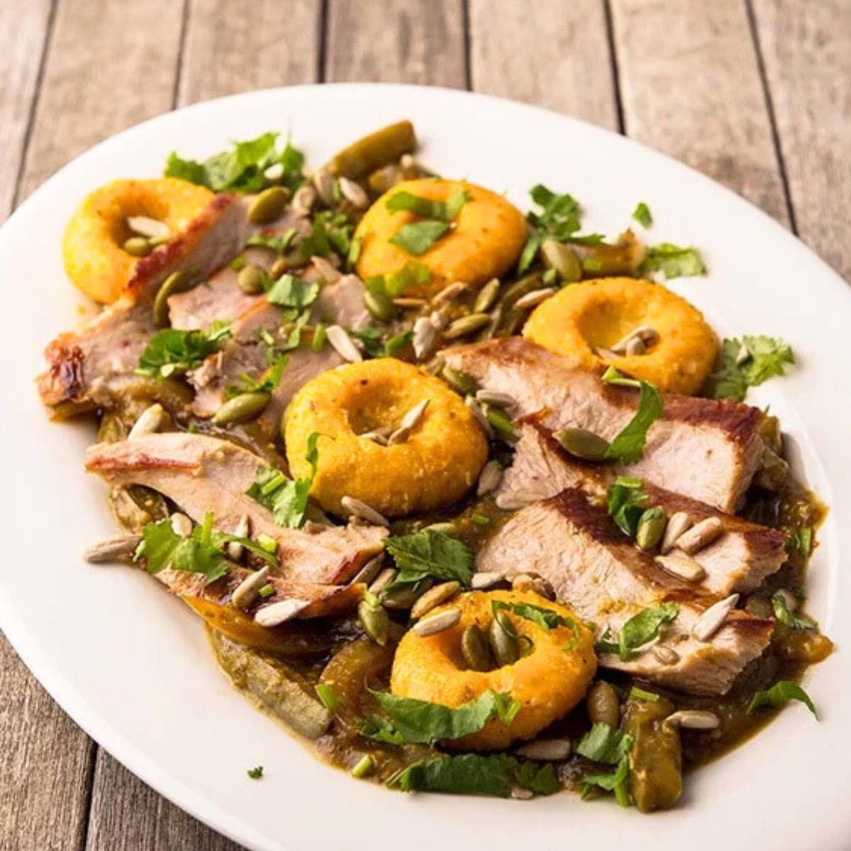 A plate of a colorful and flavorful dish. The dish features slices of roasted pheasant, topped with pumpkin dumplings, a green sauce, and pumpkin seeds. Fresh herbs garnish the dish.