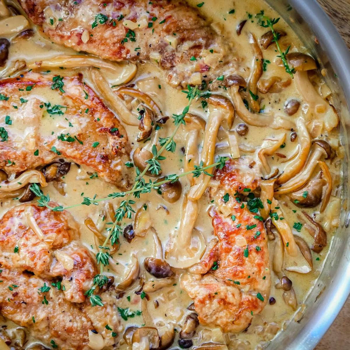 A skillet filled with a delicious-looking mushroom and pheasant dish. The chicken is golden brown and tender, and it's covered in a creamy sauce with plenty of mushrooms. A sprig of thyme adds a touch of freshness.