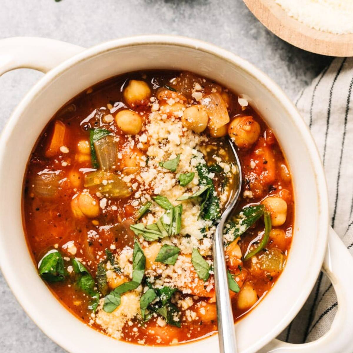 A white bowl filled with a tomato-based chickpea soup. The soup is topped with grated Parmesan cheese, fresh basil, and a spoon.