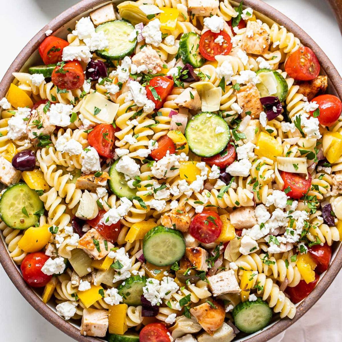 A bowl of colorful Greek pasta salad with spiral pasta, cherry tomatoes, cucumbers, red onion, artichoke hearts, olives, feta cheese, and grilled chicken.