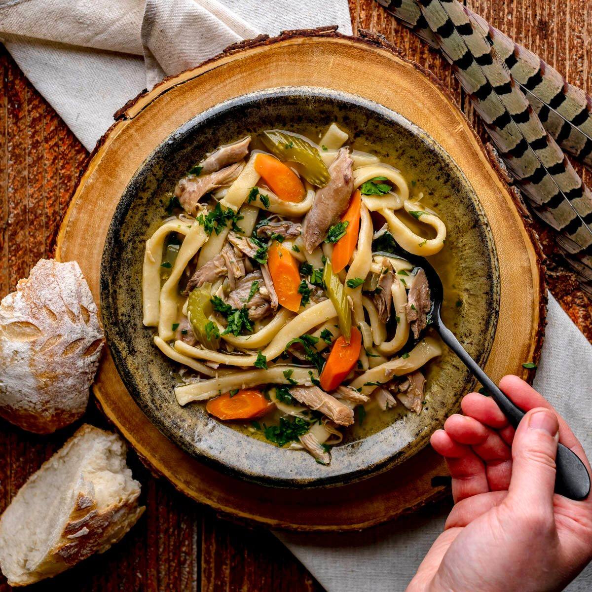 A bowl of hearty pheasant noodle soup. The soup is filled with tender meat, carrots, celery, and egg noodles. A hand is holding a spoon, ready to take a bite.