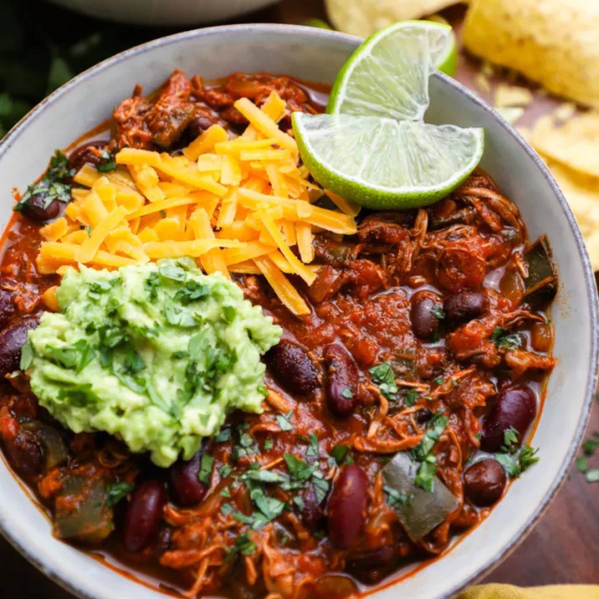 A bowl of hearty chili topped with shredded cheese, guacamole, lime wedges, and cilantro.