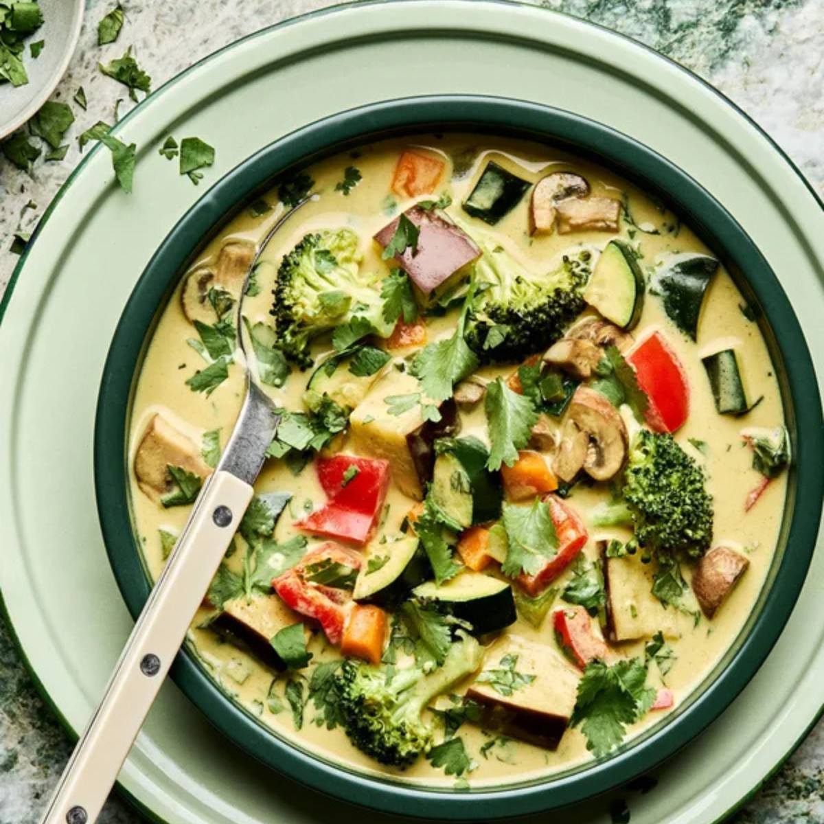 A green bowl filled with a vibrant vegetable curry. The curry is made with tofu, broccoli, zucchini, bell peppers, and mushrooms, all simmered in a creamy sauce. Fresh cilantro is sprinkled on top.