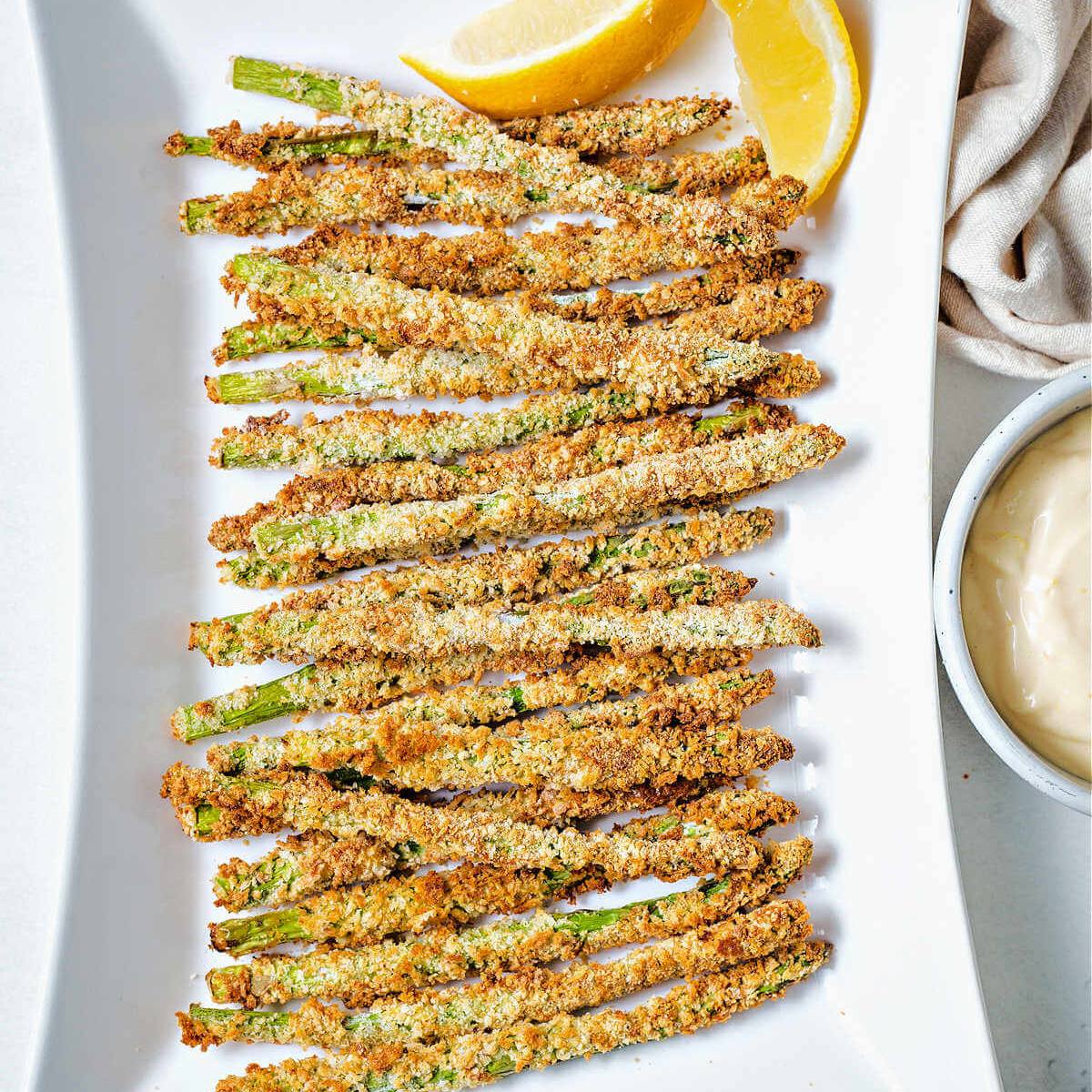 A plate of crispy, golden-brown asparagus fries with lemon wedges and a side of dipping sauce.