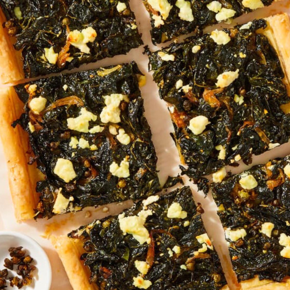 A close-up of a rectangular tart filled with sautéed kale, onions, and feta cheese, cut into squares.